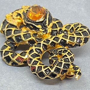 May include: A gold-toned snake brooch with black and gold detailing. The snake's body is covered in a geometric pattern, and it has a large, amber-colored stone for an eye. The brooch is a vintage accessory.