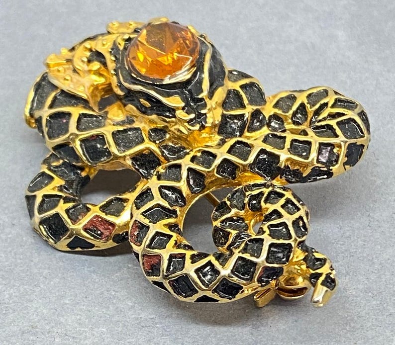 May include: A gold-toned snake brooch with black and gold detailing. The snake's body is covered in a geometric pattern, and it has a large, amber-colored stone for an eye. The brooch is a vintage accessory.
