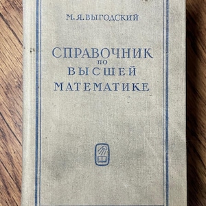 May include: A vintage book with a light gray cover and blue text. The title, in Russian, reads "Handbook of Higher Mathematics." The book has a rectangular shape and is set against a wooden background.