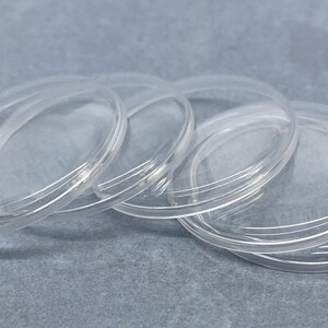 May include: A collection of five clear, circular, flat plastic discs with smooth, raised edges. These transparent, round components are arranged in a slightly overlapping, fanned-out formation, showcasing their glossy, see-through material quality.