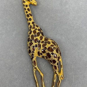 K&ouml;nnte beinhalten: Goldfarbene Giraffenbrosche mit braunen und bernsteinfarbenen Flecken. Die Giraffe hat einen langen Hals und Beine, mit einem kleinen Kristallakzent auf dem Kopf. Die Brosche ist ein dekoratives Accessoire.
