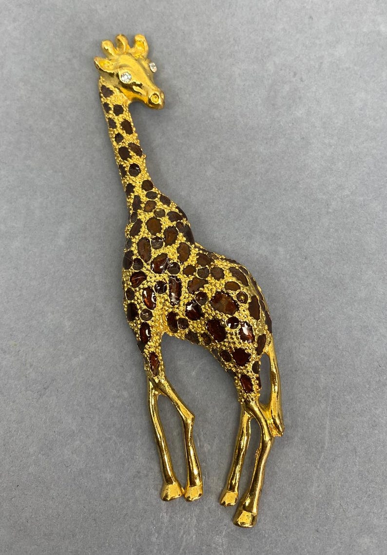 K&ouml;nnte beinhalten: Goldfarbene Giraffenbrosche mit braunen und bernsteinfarbenen Flecken. Die Giraffe hat einen langen Hals und Beine, mit einem kleinen Kristallakzent auf dem Kopf. Die Brosche ist ein dekoratives Accessoire.