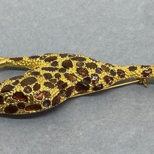 K&ouml;nnte beinhalten: Eine goldfarbene Giraffenbrosche mit braunen und bernsteinfarbenen Akzenten. Der K&ouml;rper der Giraffe ist mit braunen und bernsteinfarbenen Steinen bedeckt, und der Kopf hat kleine, klare Kristalle. Die Brosche liegt auf einer grauen Oberfl&auml;che.