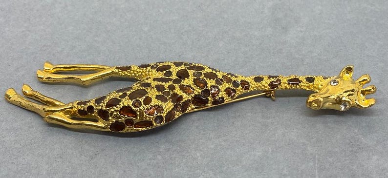 K&ouml;nnte beinhalten: Eine goldfarbene Giraffenbrosche mit braunen und bernsteinfarbenen Akzenten. Der K&ouml;rper der Giraffe ist mit braunen und bernsteinfarbenen Steinen bedeckt, und der Kopf hat kleine, klare Kristalle. Die Brosche liegt auf einer grauen Oberfl&auml;che.