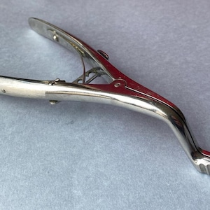 K&ouml;nnte beinhalten: Ein silberfarbenes Werkzeug im Vintage-Stil mit einem gebogenen Griff und einem scharnierartigen, klammerartigen Kopf. Das Werkzeug hat einen strukturierten Griff am Ende des Griffs, wahrscheinlich aus Metall.