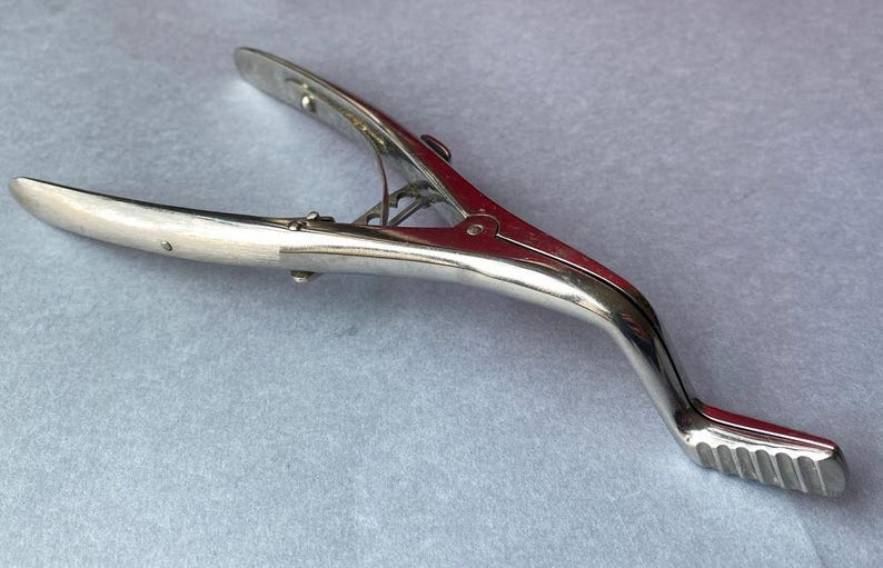 K&ouml;nnte beinhalten: Ein silberfarbenes Werkzeug im Vintage-Stil mit einem gebogenen Griff und einem scharnierartigen, klammerartigen Kopf. Das Werkzeug hat einen strukturierten Griff am Ende des Griffs, wahrscheinlich aus Metall.