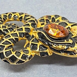 May include: A gold-toned snake brooch with black enamel detailing and a large amber-colored gemstone. The snake is coiled, with intricate scale patterns and a pin closure on the back. A unique jewelry piece.