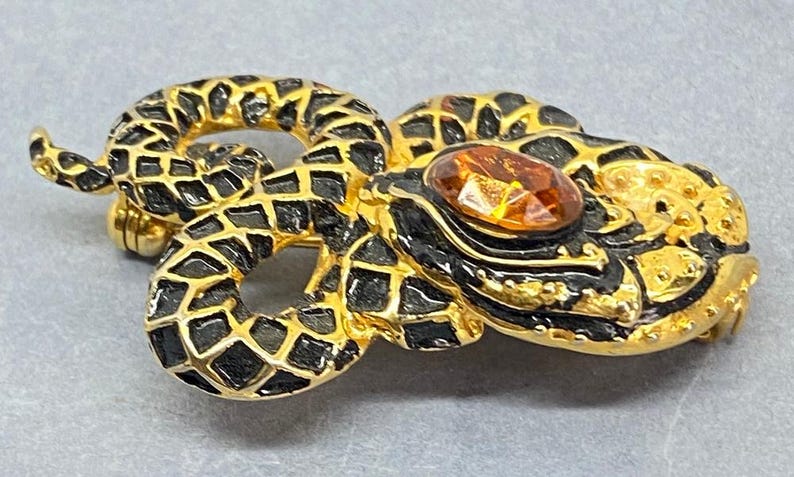 May include: A gold-toned snake brooch with black enamel detailing and a large amber-colored gemstone. The snake is coiled, with intricate scale patterns and a pin closure on the back. A unique jewelry piece.