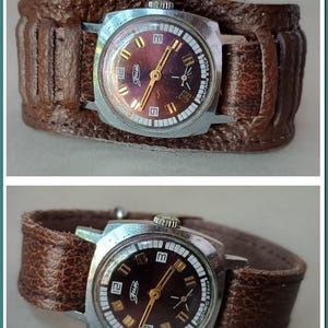 Puede incluir: Un reloj de pulsera vintage con una esfera cuadrada y una correa de cuero marrón. La esfera del reloj tiene un fondo morado con marcadores de hora dorados y una aguja de segundos plateada. El reloj está etiquetado como "Zarya" y "Сделано в СССР".
