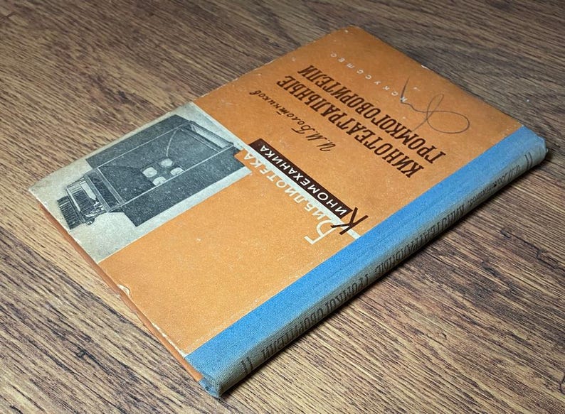 May include: A vintage book with an orange cover and blue spine, titled in Russian. The cover features an illustration of a film projector. The book's title is "Kinoteatralnye Gromkogovoriteli" and the author is listed as "M. Bogomolov".