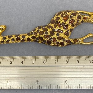 K&ouml;nnte beinhalten: Goldfarbene Giraffenbrosche mit braunen Flecken und bernsteinfarbenen Akzenten. Die Giraffe liegt, die Beine ausgestreckt. Die Brosche misst etwa 12 cm in der L&auml;nge.
