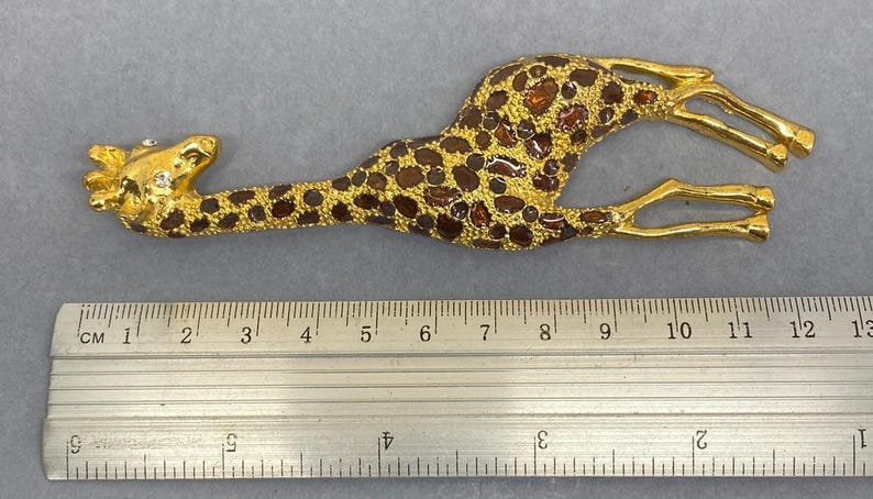 K&ouml;nnte beinhalten: Goldfarbene Giraffenbrosche mit braunen Flecken und bernsteinfarbenen Akzenten. Die Giraffe liegt, die Beine ausgestreckt. Die Brosche misst etwa 12 cm in der L&auml;nge.