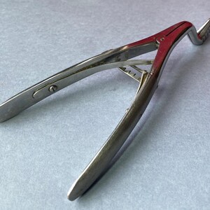 K&ouml;nnte beinhalten: Ein chirurgisches Vintage-Instrument, wahrscheinlich eine Zange oder Klammer, aus poliertem Metall. Das Werkzeug hat ein gebogenes Design mit einem roten Akzent am zentralen Gelenk und einem gezahnten Ende. Das Instrument ist zum Greifen oder Halten bestimmt.