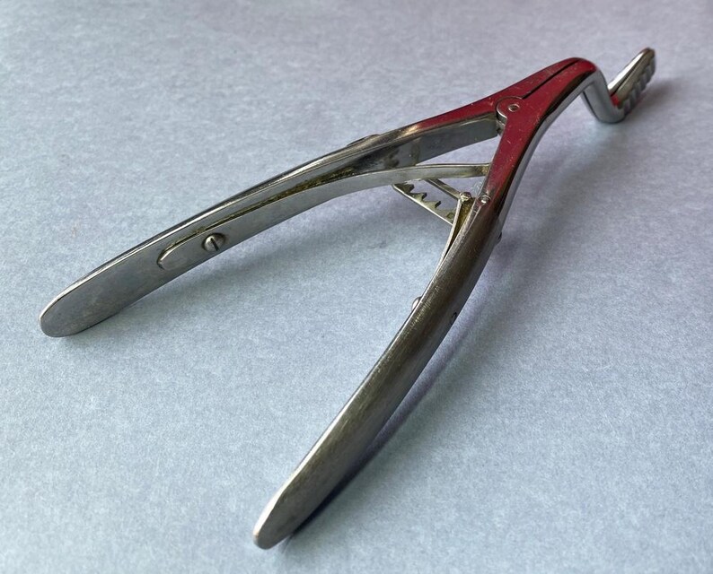 K&ouml;nnte beinhalten: Ein chirurgisches Vintage-Instrument, wahrscheinlich eine Zange oder Klammer, aus poliertem Metall. Das Werkzeug hat ein gebogenes Design mit einem roten Akzent am zentralen Gelenk und einem gezahnten Ende. Das Instrument ist zum Greifen oder Halten bestimmt.