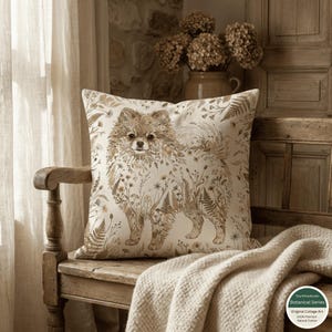 May include: A cushion with a Pomeranian dog design, set on a wooden bench. The dog is illustrated with botanical elements in brown and beige tones. The cushion is placed on a bench with a cream-coloured blanket, creating a cosy, rustic feel.