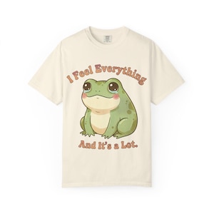 May include: Cream-colored t-shirt featuring a cartoon frog with large eyes and rosy cheeks. The frog is light green with darker green spots. Text above and below the frog reads "I Feel Everything" and "And It's a Lot."