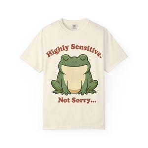 Könnte beinhalten: Cremefarbenes T-Shirt mit einem Cartoon-Frosch und dem Text "Highly Sensitive. Not Sorry..." in rötlich-brauner Schrift. Der Frosch ist grün mit cremefarbenem Bauch und geschlossenen Augen. Das T-Shirt hat kurze Ärmel und ist aus weichem Material.