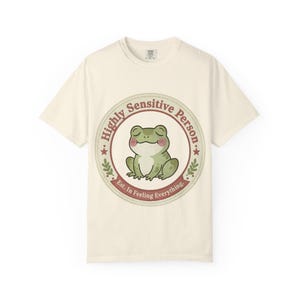 May include: Cream-colored t-shirt featuring a circular graphic with a green frog illustration. The graphic includes the text "Highly Sensitive Person" and "Est. In Feeling Everything."