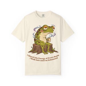 Könnte beinhalten: Cremefarbenes T-Shirt mit einer Cartoon-Frosch, der auf einem Baumstumpf sitzt und eine Tasse hält. Der Frosch ist grün mit braunen Flecken und hat einen müden Ausdruck. Der Text auf dem Shirt lautet: "I Absorb the Energy of Every Room I Walk Into and I'm Exhausted."
