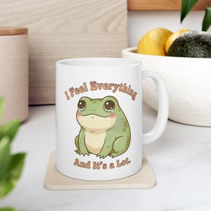 May include: White ceramic mug featuring a cartoon frog with the text "I Feel Everything And It's a Lot." The frog is green with a light green belly and large, expressive eyes. The text is in a warm, brown color.