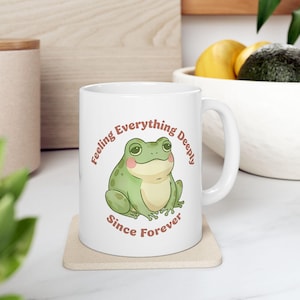May include: White ceramic mug featuring a cartoon frog with rosy cheeks and the text "Feeling Everything Deeply Since Forever" in a circular design. The frog is green with a cream-colored belly.
