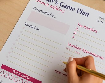 May include: A printed planner titled "Today's Game Plan (Mom's Edition)" with sections for gratitude, to-do lists, and appointments. A hand is writing with a gold pen. Black glasses and a white mug are also visible.