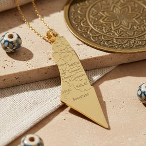 May include: A gold-colored pendant necklace in the shape of a map of Palestine, with city names engraved. The necklace hangs on a delicate chain. Decorative beads and a brass plate are in the background.