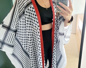 May include: A black and white patterned kimono with a red trim. The kimono is draped over a black top and leggings. The pattern is a repeating geometric design. A gold necklace is visible.