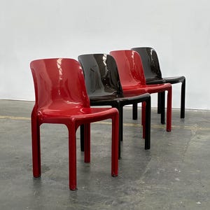 Op de afbeelding: Vier moderne stoelen op een rij. De stoelen zijn gemaakt van glanzend plastic. Twee zijn rood, één is donkerbruin en één is zwart. De stoelen hebben een eenvoudig, minimalistisch ontwerp met rechte poten en een gebogen rug.
