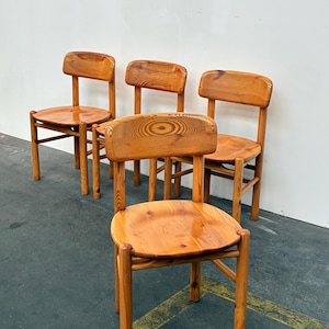 Può includere: Quattro sedie in legno con finitura naturale sono disposte in una stanza. Le sedie hanno sedili arrotondati, gambe dritte e schienale. La venatura del legno è visibile sui sedili e sugli schienali. Le sedie sono poste contro un muro bianco.
