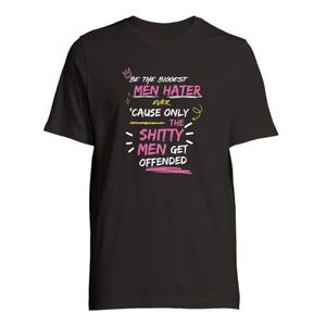 Puede incluir: Una camiseta negra de manga corta con texto en blanco y rosa que dice: BE THE BIGGEST MEN HATER EVER 'CAUSE ONLY THE SHITTY MEN GET OFFENDED. El diseño incluye un pequeño icono de corona rosa y líneas y flechas decorativas de color amarillo.