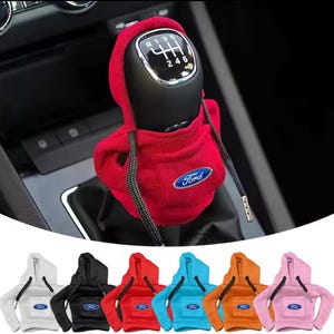 Peut inclure: Un couvre-levier de vitesse rouge en forme de sweat à capuche avec le logo Ford, présenté sur le levier de vitesse d'une voiture. Des housses supplémentaires en blanc, noir, rouge, bleu, orange et rose sont affichées en dessous. Les housses ont des cordons de serrage et le logo Ford.