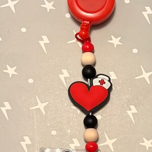 May include: A red retractable badge reel with a heart-shaped charm featuring a nurse's cap. The charm is attached to a string of black, beige, and red beads. The reel has a clear plastic badge holder.