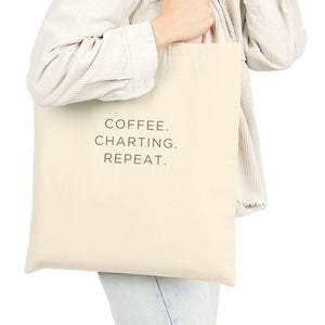 May include: A beige tote bag with the words "COFFEE. CHARTING. REPEAT." printed in black. The bag is held by a person wearing a white jacket and blue jeans.