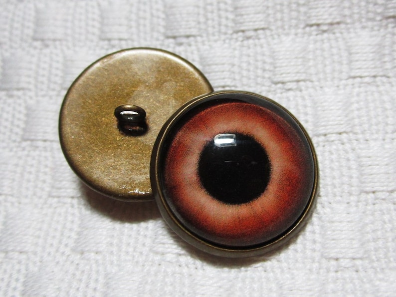 Peut inclure: Deux boutons ronds, l'un avec une finition en laiton et l'autre avec un motif d'&oelig;il rouge et noir.