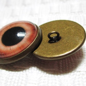 Peut inclure: Deux boutons ronds en bronze avec un motif d'&oelig;il rouge et noir. Le bouton de gauche a une pupille noire et un iris rouge. Le bouton de droite est le dos du bouton et a une boucle pour coudre.
