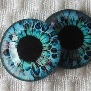 May include: Two round glass eye cabochons with a detailed iris design. The irises are a mix of teal, turquoise, and blue, with a black pupil. The cabochons are set against a white textured background.