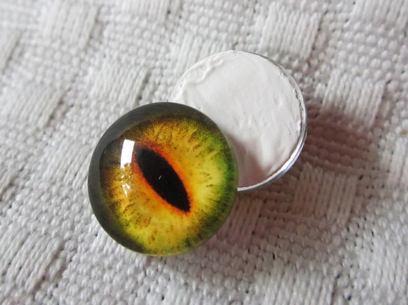 May include: A close-up of a yellow and green glass eye with a black pupil. The eye is sitting on a white surface.