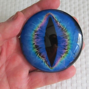 May include: A round, glass eye with a blue iris and a black pupil. The iris has a detailed pattern with hints of purple, green, and yellow. The eye is held in a hand.