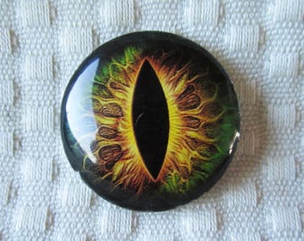 May include: A round button with a green, yellow, and black eye design. The eye is a vertical oval shape with a black pupil and a yellow iris with green flames.