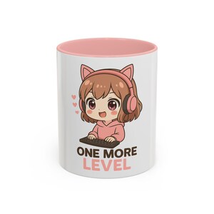 Könnte beinhalten: Weißer Keramikbecher mit rosa Innenraum. Der Becher zeigt eine Cartoon-Illustration eines Mädchens mit braunen Haaren, das Kopfhörer und einen rosa Kapuzenpullover trägt und eine Tastatur hält. Der Text "ONE MORE LEVEL" ist darunter gedruckt.