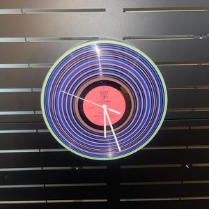 May include: A round wall clock made from a vinyl record. The clock face features a pink center with white hands and a light green border. The record has blue, purple, and pink concentric circles.