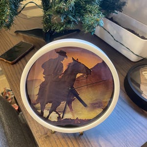 May include: A round clock with a white frame and gold-colored legs. The clock face features a silhouette of a cowboy riding a horse against a sunset backdrop. The clock hands are black.
