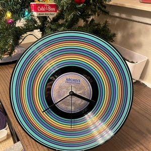 May include: A vinyl record clock with a colourful striped design in shades of blue, yellow, and pink. The clock face features a vintage record label and black clock hands. The clock is displayed on a wooden surface.