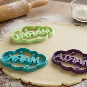 May include: Three cloud-shaped cookie cutters spelling out the names "Mark", "Tara", and "Marta". The cutters are in green, teal, and purple. The cutters are on a rolled-out dough with a rolling pin and a bowl of flour in the background.