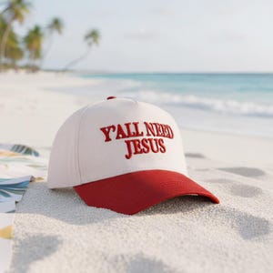 May include: A white baseball cap with a red brim and a red button on top. The phrase "Y'ALL NEED JESUS" is embroidered in red on the front of the cap. The cap is resting on a sandy beach with the ocean in the background.