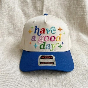 May include: A white baseball cap with a blue brim. The cap features the embroidered phrase "have a good day" in colorful letters and star accents. A product tag is visible on the brim.