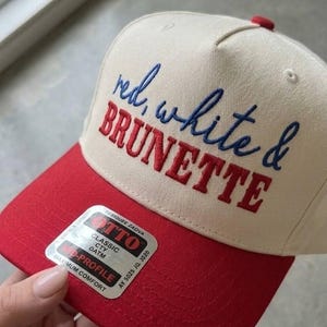 Peut inclure: Casquette de baseball avec une visi&egrave;re et un bouton rouges, et une couronne cr&egrave;me. Le chapeau porte la broderie "red, white & BRUNETTE" en rouge et bleu. Un autocollant est visible sur la visi&egrave;re.