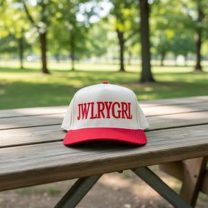 May include: A white baseball cap with a red brim and the text "JWLRYGRL" in red block letters. The hat is resting on a wooden picnic table in a park setting.
