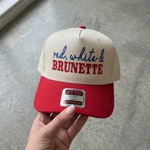 Peut inclure: Casquette de baseball avec une visi&egrave;re rouge et une couronne cr&egrave;me. Le texte "red, white & BRUNETTE" est brod&eacute; sur le devant en bleu et rouge. La casquette a un bouton rouge sur le dessus et une &eacute;tiquette sur la visi&egrave;re.